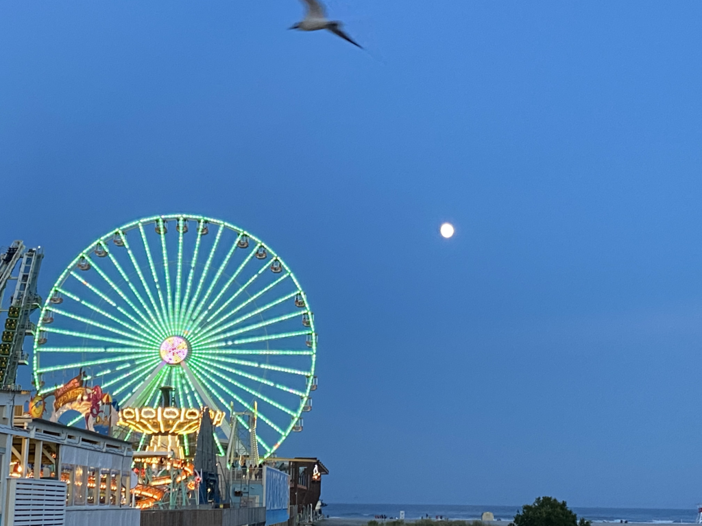 an illuminated Ferris wheel shines in the late summer sky with the full moon glowing in the distance